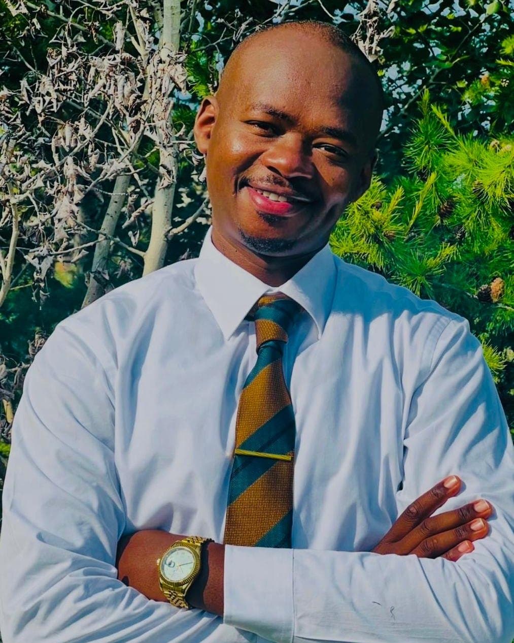 David Nyakundi Ombasa, arms folded, smiling, standing outdoors in front of trees