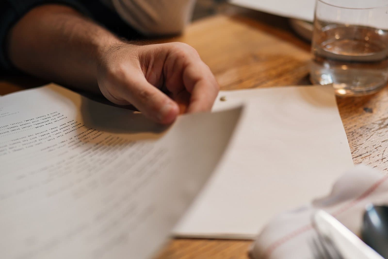 A person's hands turning a printed page on a wooden table beside a glass of water.