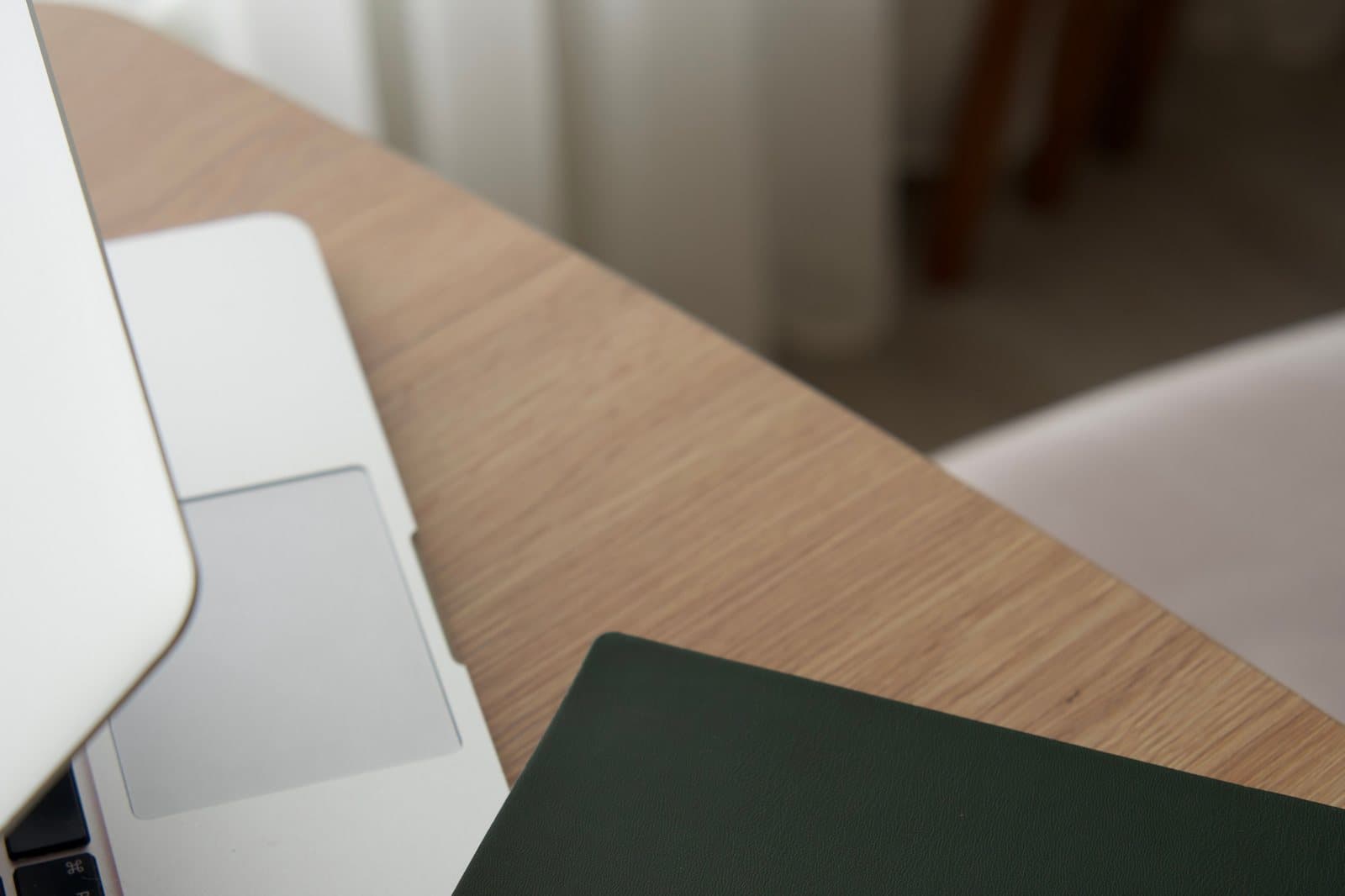 A laptop and a closed notebook on the corner of a light wooden table.