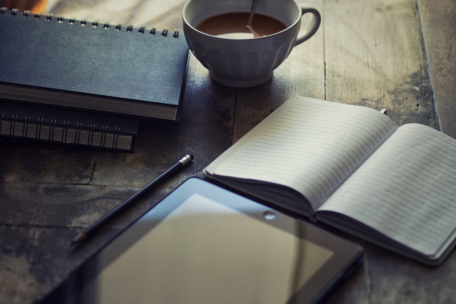 An open notebook, a tablet and a cup of coffee on a dark wooden table.