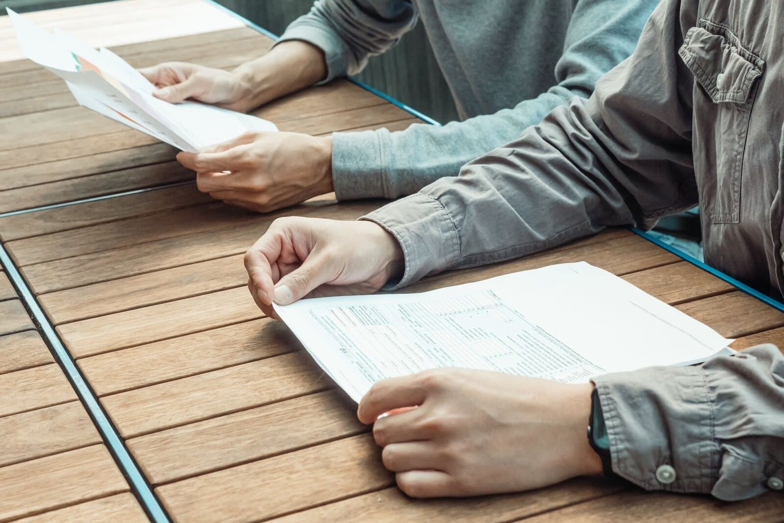 Two people's hands going through printed forms on a wooden table.
