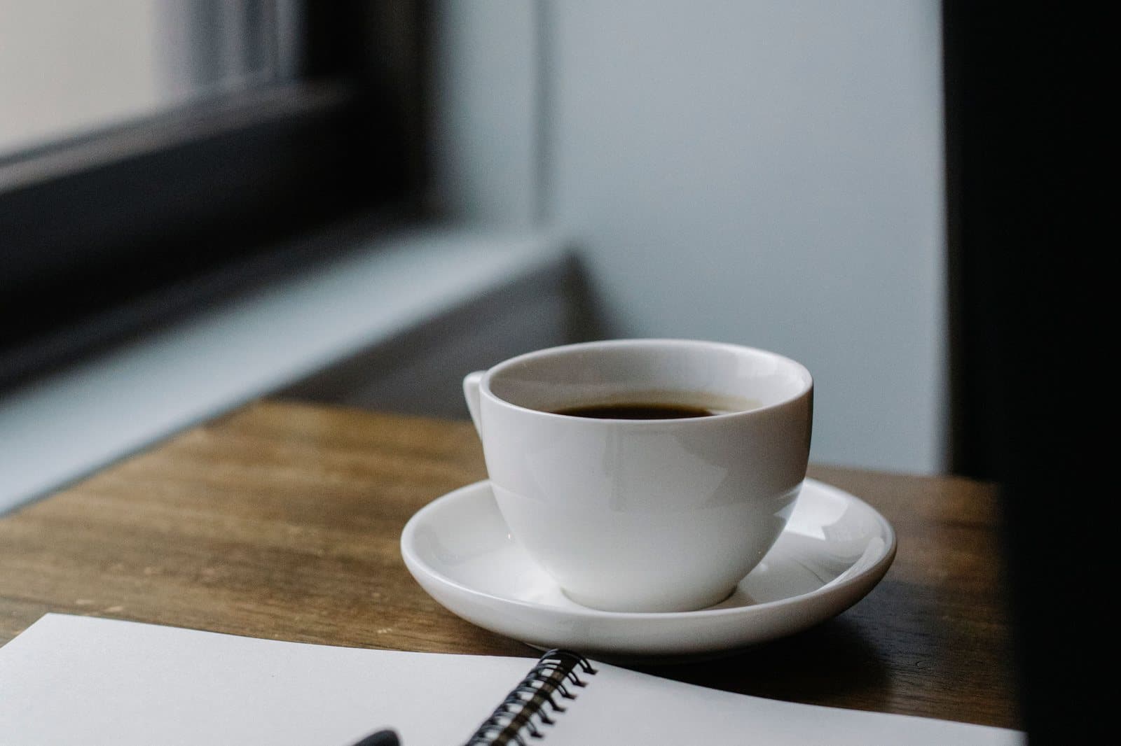 A cup of coffee and a closed notebook on a table beside a window.