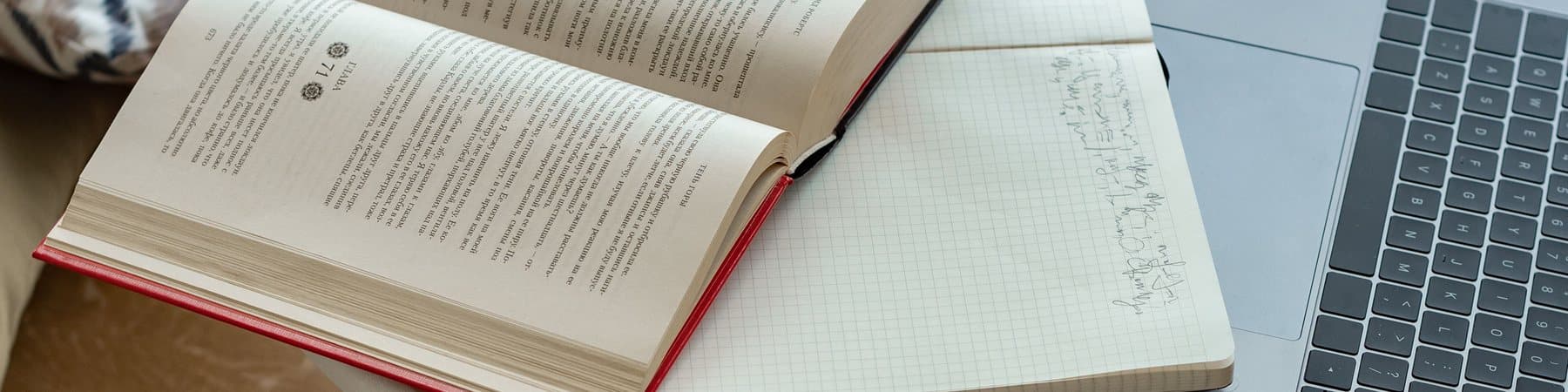 An open book and a laptop side by side on a table beside a window.