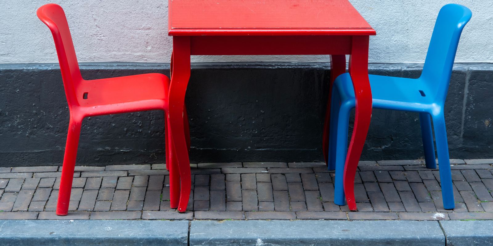 A child-sized red table with red and blue chairs, empty, on a paved street.