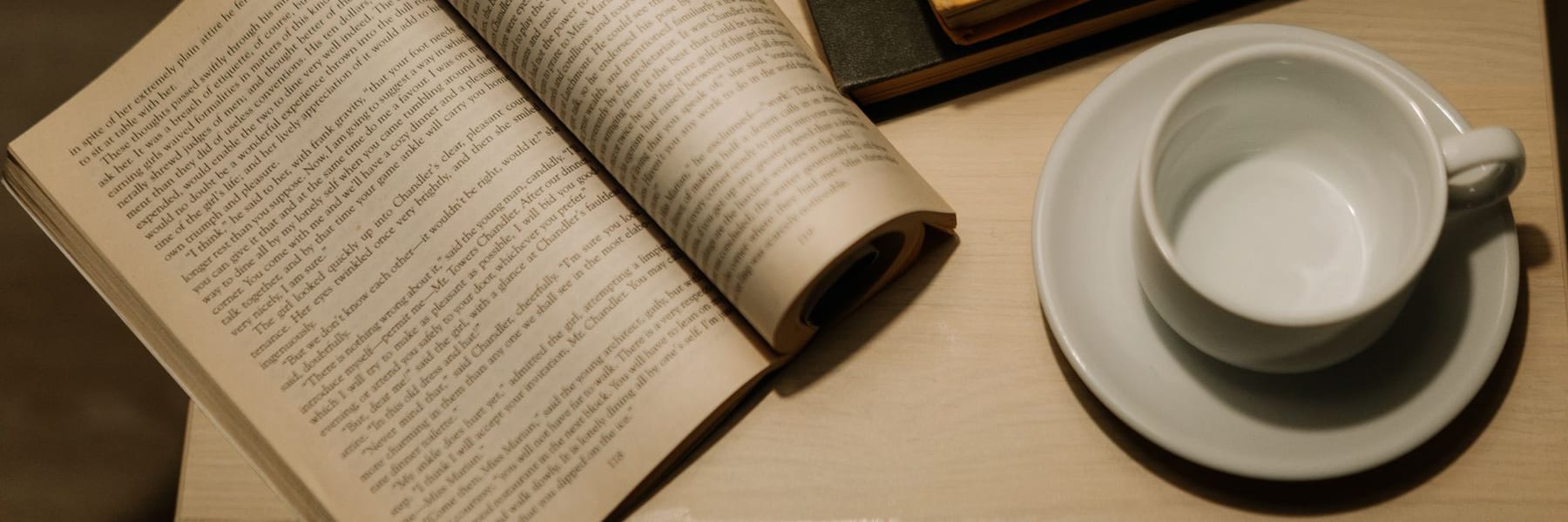 An open book beside a cup and saucer on a wooden table.