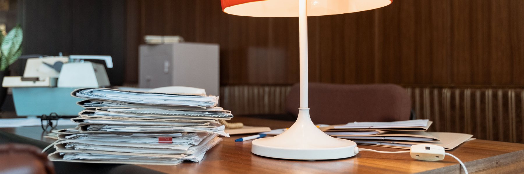 Stacks of paperwork and a lit lamp on a wooden desk.
