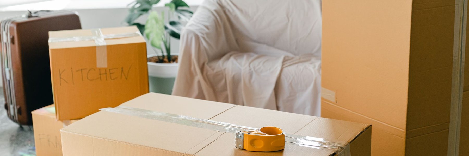 Packing boxes, one marked Kitchen, stacked beside an armchair in a bright room.